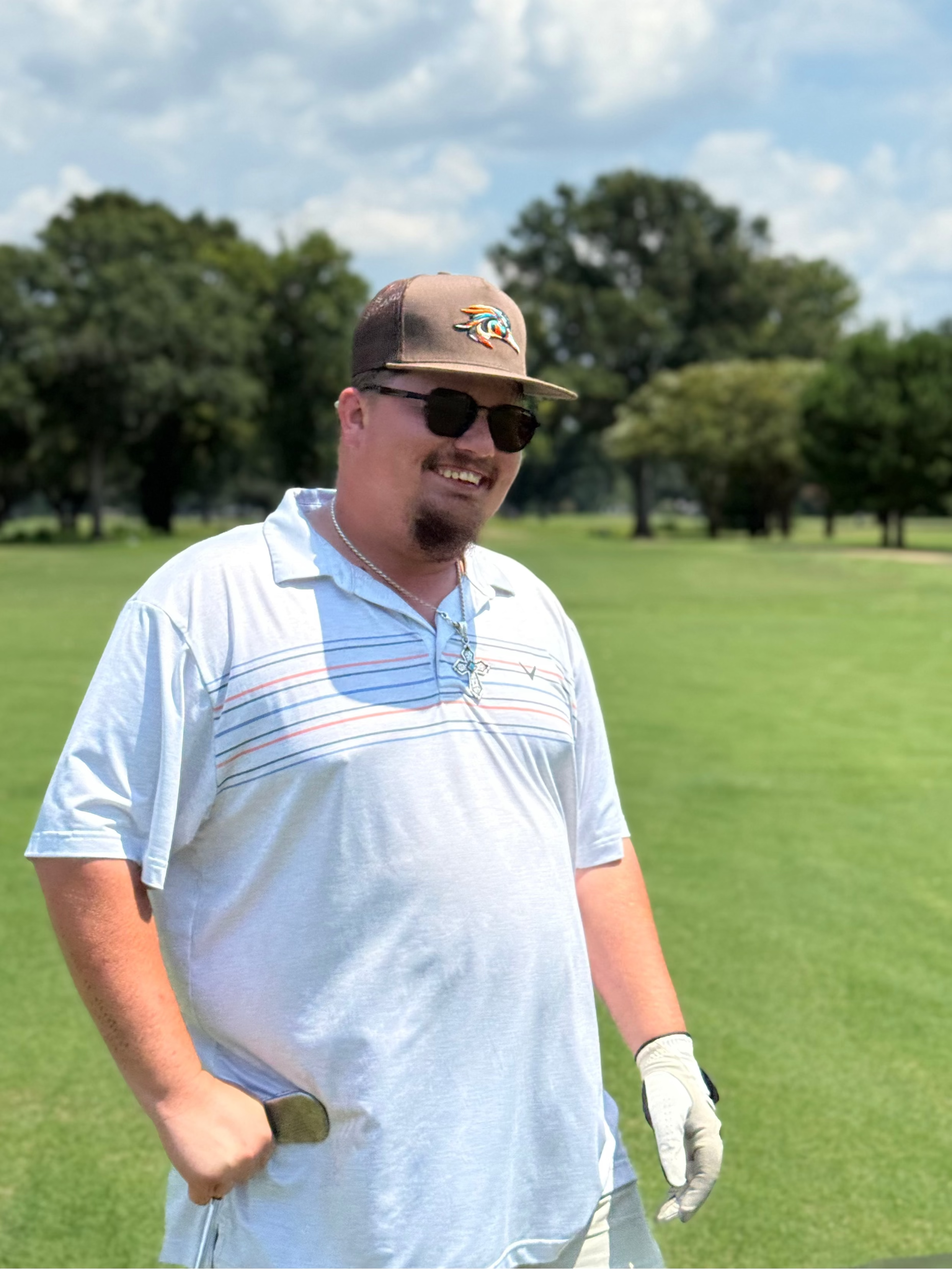 Man on a golf course wearing sunglasses and a cap with trees in the background